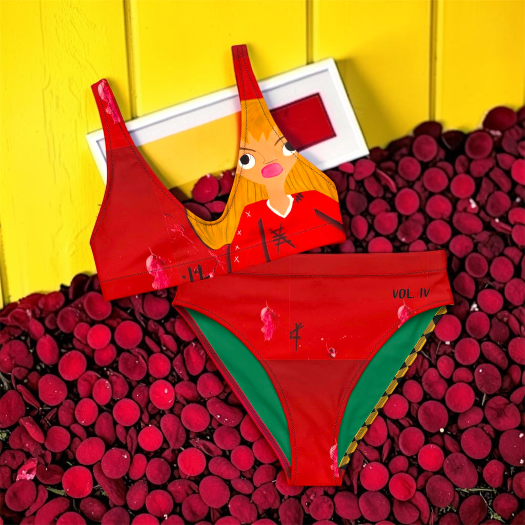 Vol IV — The Doll Wall Goddess Bikini in scarlet red — top featuring golden lady Doll Wall print, bottom with Vol IV detail and forest green lining, styled on bed of red rose petals against yellow wall — Golden Expression Art Limited Edition
