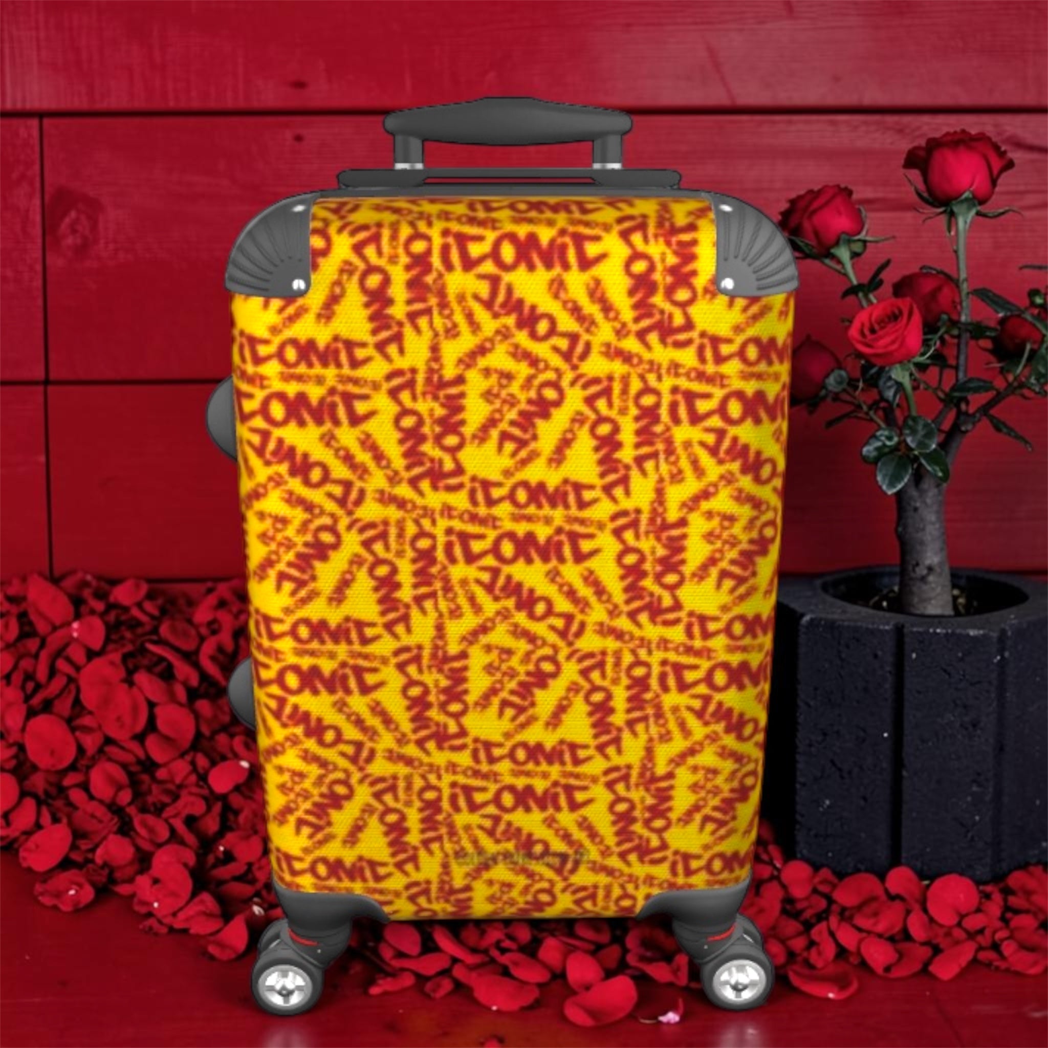 The ICONIC Getaway carry-on luggage — front view, all-over ICONIC print in golden yellow and scarlet red, spinner wheels, red wall backdrop