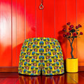 Vol II — The Sacred Garden Short, front view, all-over Eden art print, Doll Wall muse repeating pattern, golden yellow base with blue and green tones, elastic waistband, red editorial backdrop, Golden Expression Art
