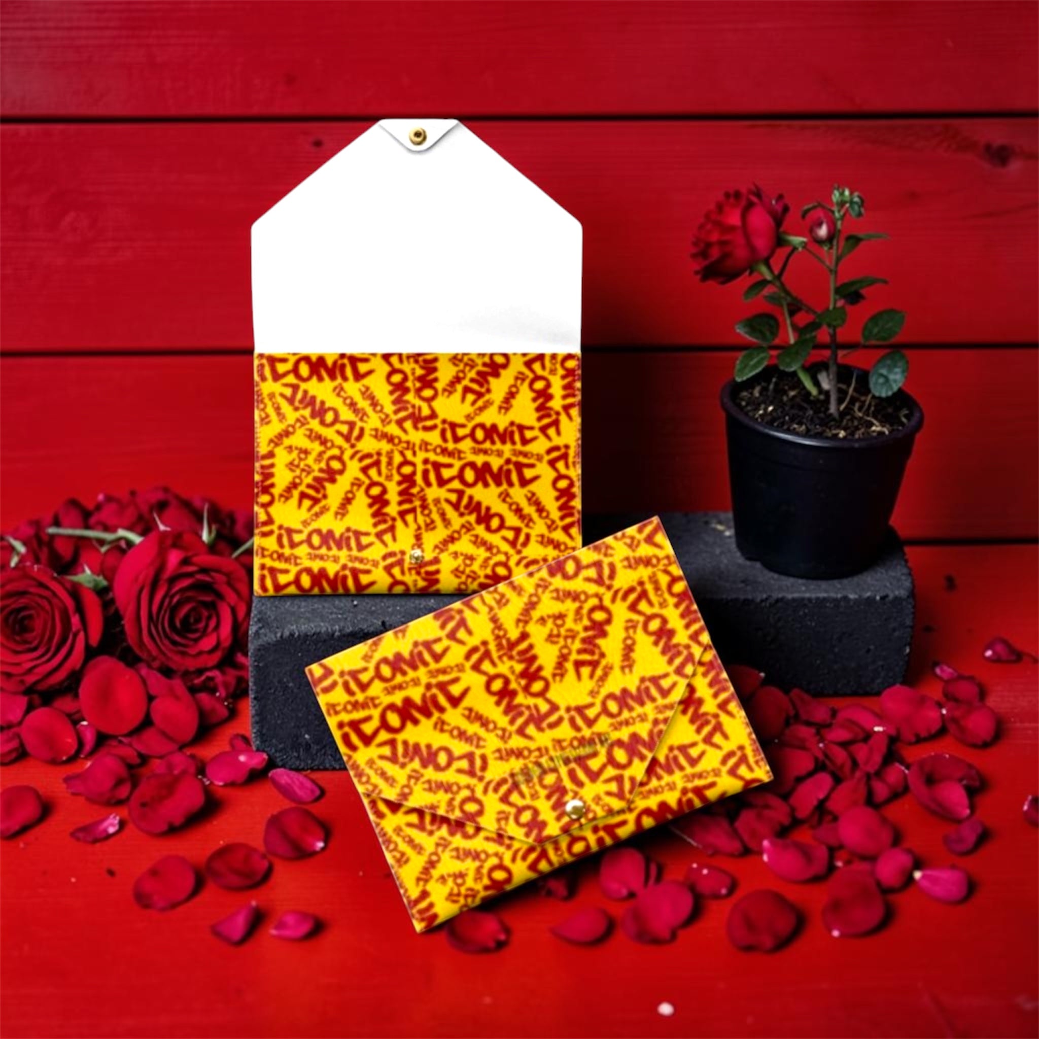 The ICONIC Envelope Card Holder — front and wallet view, golden yellow ICONIC all-over print with scarlet red lettering, gold snap closure, rose petal editorial backdrop