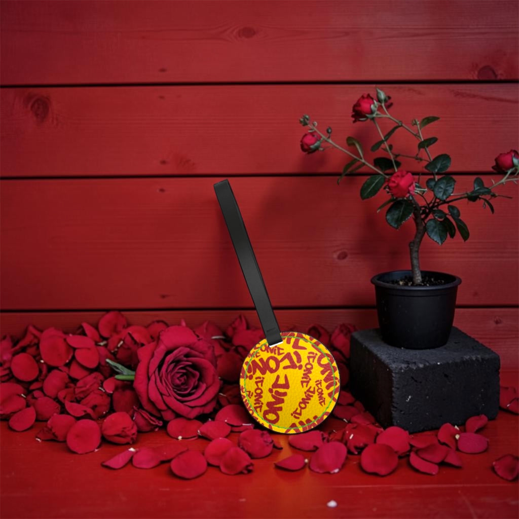 Vol IV — The ICONIC Charm — round leather charm with all-over scarlet red ICONIC lettering on golden yellow, black wrist strap, rose petal backdrop