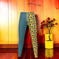 Vol II — The Eden Two-World Jogger, front view, split color-block, left leg solid teal, right leg all-over Eden repeat print animated woman golden yellow base blue and green tones, white drawstring waistband, GEA